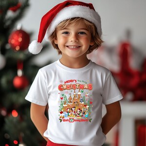 May include: A white t-shirt featuring a colorful graphic of Disney characters and a castle, with the text "Mickey's Very Merry Christmas" and "Walt Disney World". The child is wearing a red and white Santa hat.