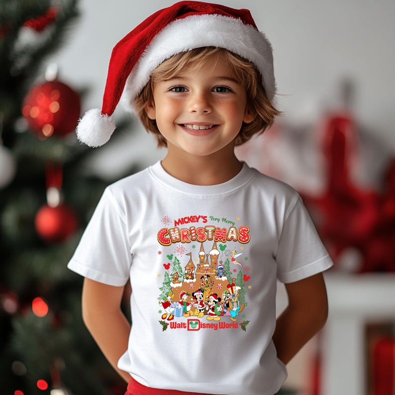 May include: A white t-shirt featuring a colorful graphic of Disney characters and a castle, with the text "Mickey's Very Merry Christmas" and "Walt Disney World". The child is wearing a red and white Santa hat.