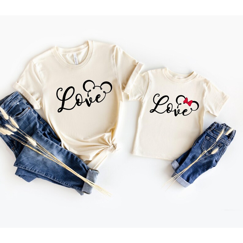 K&ouml;nnte beinhalten: Zwei beige T-Shirts mit dem Wort "Love" in schwarzer Schrift. Das gr&ouml;&szlig;ere Shirt hat eine schwarze Mickey-Maus-Silhouette. Das kleinere Shirt hat eine schwarze Mickey-Maus-Silhouette mit einer roten Schleife.