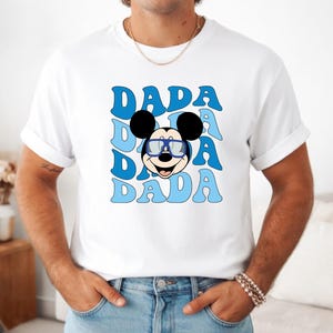 K&ouml;nnte beinhalten: Wei&szlig;es T-Shirt mit einem Cartoon-Mickey-Mouse-Design mit Brille und dem Wort "DADA" in Blau, dreimal wiederholt. Die Schriftart ist retro und wellenf&ouml;rmig. Das T-Shirt wird von einer Person getragen.