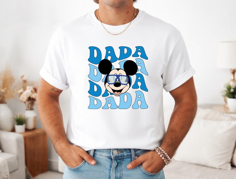 K&ouml;nnte beinhalten: Wei&szlig;es T-Shirt mit einem Cartoon-Mickey-Mouse-Design mit Brille und dem Wort "DADA" in Blau, dreimal wiederholt. Die Schriftart ist retro und wellenf&ouml;rmig. Das T-Shirt wird von einer Person getragen.