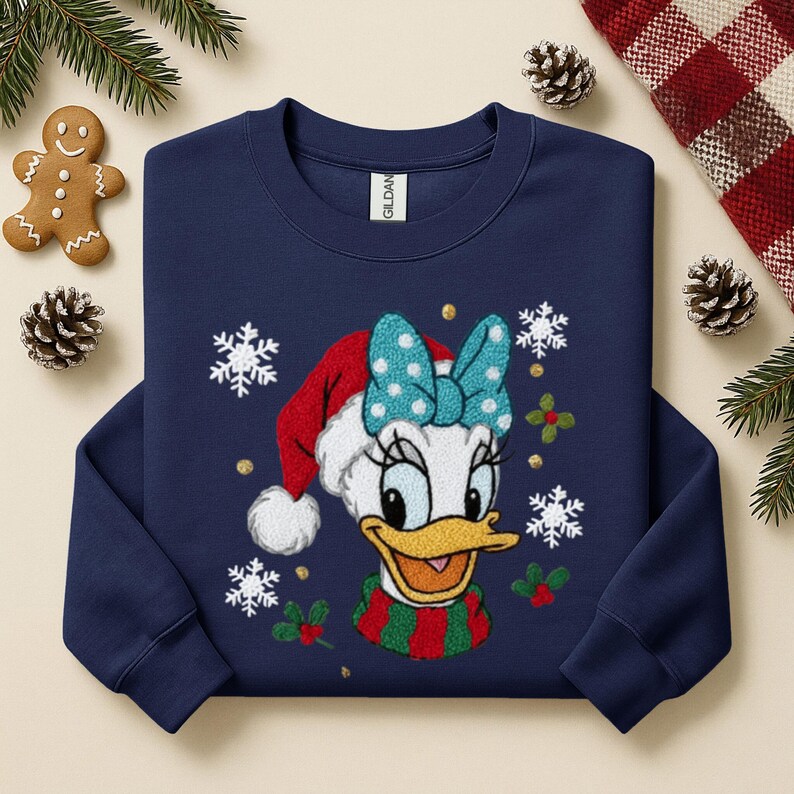 May include: Navy blue sweatshirt featuring a festive Daisy Duck design. Daisy is wearing a Santa hat with a blue bow, a striped scarf, and surrounded by snowflakes and holly. A gingerbread man and pinecones are in the background.