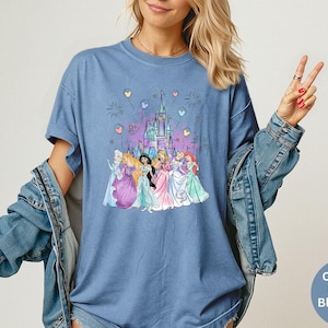 K&ouml;nnte beinhalten: Blaues Jeans-T-Shirt mit einer Grafik eines Schlosses und mehrerer Prinzessinnen. Die Grafik enth&auml;lt Feuerwerk und Ballons. Das Shirt hat eine Komfortfarbe und tr&auml;gt die Worte "Comfort Colors Blue Jean" unten rechts.
