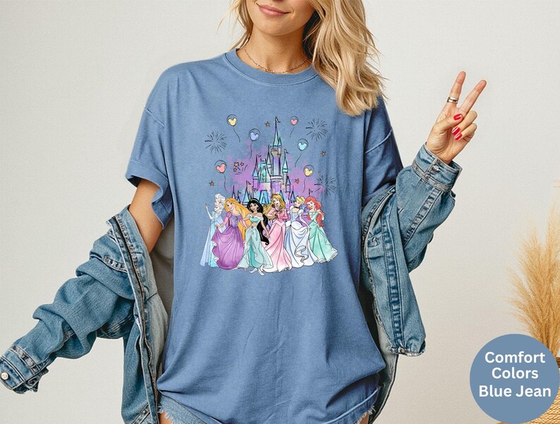 K&ouml;nnte beinhalten: Blaues Jeans-T-Shirt mit einer Grafik eines Schlosses und mehrerer Prinzessinnen. Die Grafik enth&auml;lt Feuerwerk und Ballons. Das Shirt hat eine Komfortfarbe und tr&auml;gt die Worte "Comfort Colors Blue Jean" unten rechts.