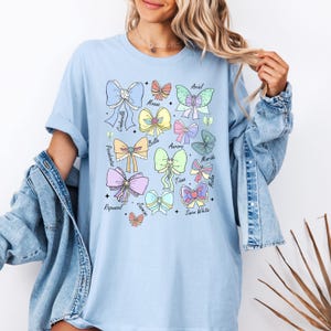 May include: Light blue t-shirt with a graphic of pastel-colored bows and the names of Disney princesses: Cinderella, Ariel, Moana, Belle, Aurora, Merida, Tiana, Mulan, Rapunzel, Jasmine, and Snow White.