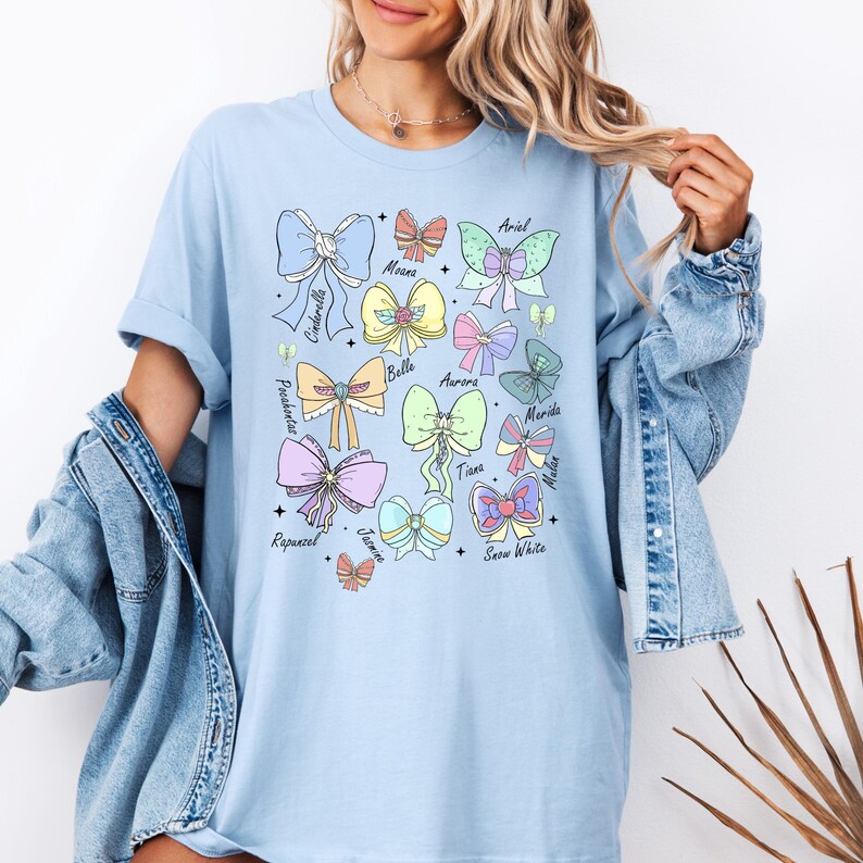 May include: Light blue t-shirt with a graphic of pastel-colored bows and the names of Disney princesses: Cinderella, Ariel, Moana, Belle, Aurora, Merida, Tiana, Mulan, Rapunzel, Jasmine, and Snow White.