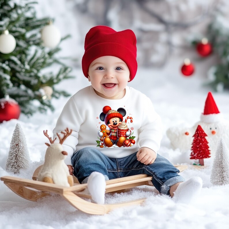 May include: A baby wearing a white long-sleeved shirt with a Christmas-themed Mickey Mouse graphic, red hat, and blue jeans, sitting on a wooden sled in a snowy setting.