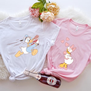 May include: Two white t-shirts featuring cartoon images of Donald Duck and Daisy Duck. The Donald Duck shirt shows him running with a bouquet of flowers. The Daisy Duck shirt shows her with hearts floating around her head.
