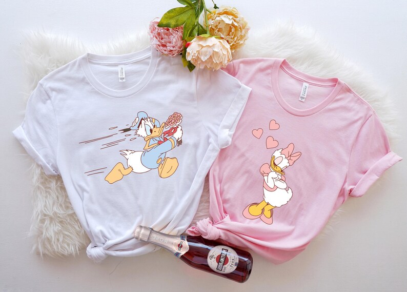 May include: Two white t-shirts featuring cartoon images of Donald Duck and Daisy Duck. The Donald Duck shirt shows him running with a bouquet of flowers. The Daisy Duck shirt shows her with hearts floating around her head.