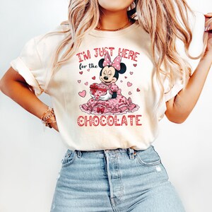 May include: Cream-colored t-shirt featuring a Minnie Mouse graphic. Minnie is dressed in a pink dress and holding a heart-shaped box. The text "I'M JUST HERE for the CHOCOLATE" is printed above and below the graphic.