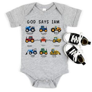 May include: A gray baby onesie with the text "God Says I Am" and illustrations of tractors. Each tractor is paired with a word describing a positive character trait, such as "handsome", "accepted", "bold", "redeemed", "protected", "courageous", "kind", "strong", and "loyal".