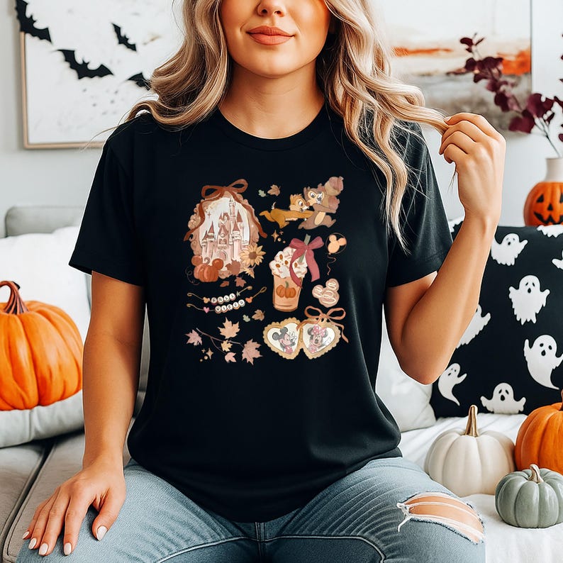 Puede incluir: Camiseta negra con un dise&ntilde;o gr&aacute;fico colorido con un castillo, Chip y Chop, un caf&eacute; con leche y Mickey y Minnie Mouse. El dise&ntilde;o tiene un tema oto&ntilde;al con calabazas y hojas.