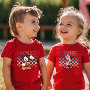 May include: Two children wearing red t-shirts with a retro checkered pattern and a graphic of Mickey Mouse and Minnie Mouse. The boy's shirt says "MICKEY" and the girl's shirt says "MINNIE".