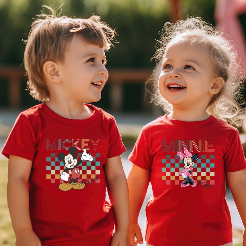 May include: Two children wearing red t-shirts with a retro checkered pattern and a graphic of Mickey Mouse and Minnie Mouse. The boy's shirt says "MICKEY" and the girl's shirt says "MINNIE".