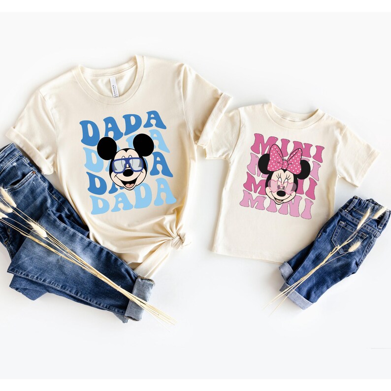 May include: Two cream coloured t-shirts with a retro design. The larger shirt has a blue and white design with Mickey Mouse wearing sunglasses and the text "DADA". The smaller shirt has a pink and white design with Minnie Mouse wearing sunglasses and the text "MINI".