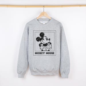 May include: A light gray sweatshirt featuring a black and white sketch of Mickey Mouse inside a square frame. The words "MICKEY MOUSE" are printed in a bold, black font below the image. The sweatshirt is hanging on a wooden hanger.