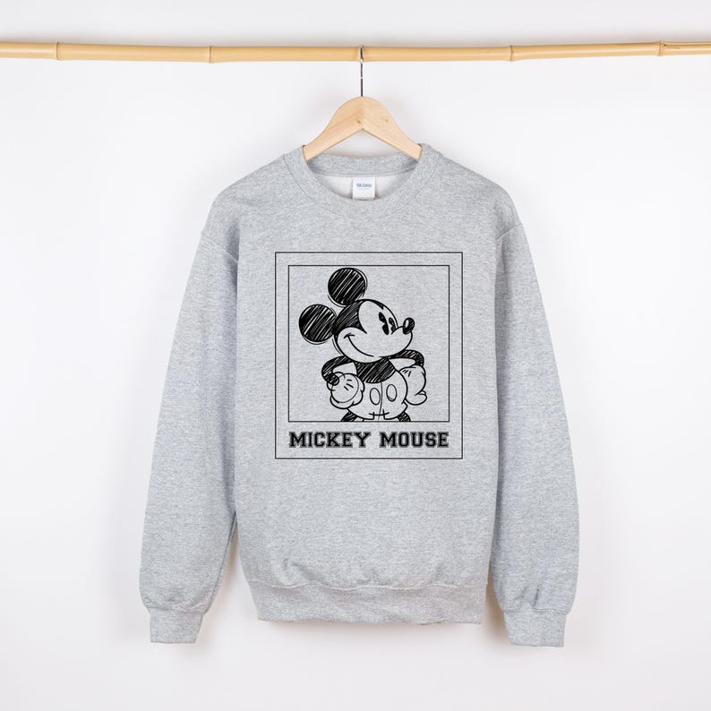 May include: A light gray sweatshirt featuring a black and white sketch of Mickey Mouse inside a square frame. The words "MICKEY MOUSE" are printed in a bold, black font below the image. The sweatshirt is hanging on a wooden hanger.