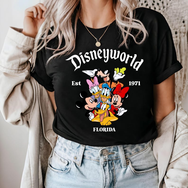 Pu&ograve; includere: Maglietta nera con una grafica bianca di Topolino, Minnie Mouse, Paperino, Pippo e Pluto. La grafica include il testo "Disneyworld Est 1971 Florida".