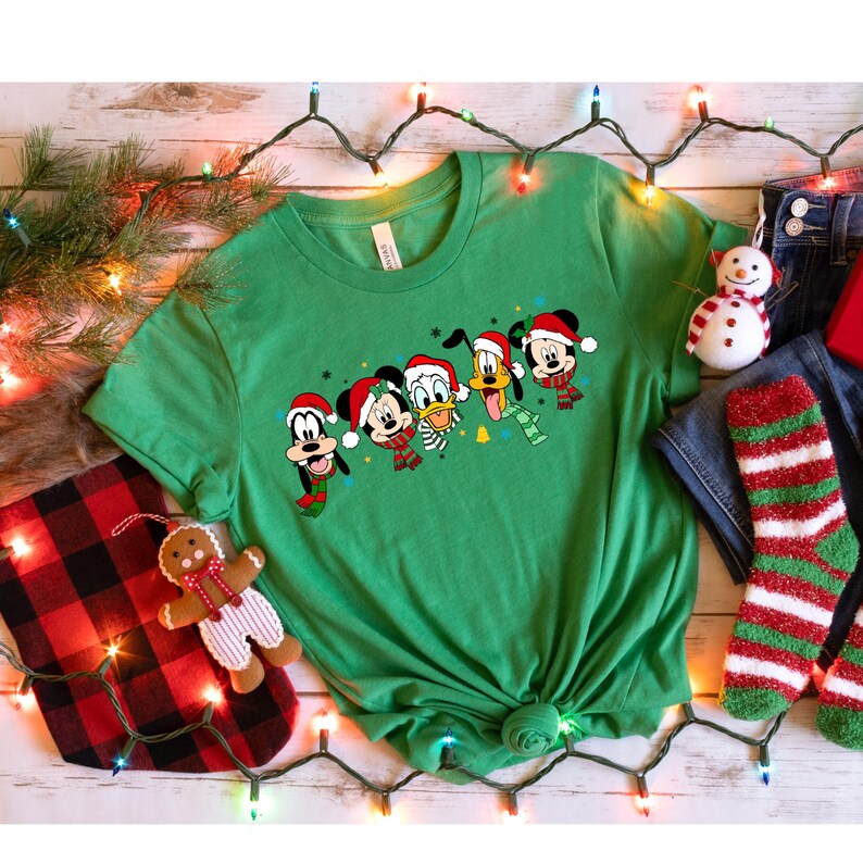 May include: Green t-shirt with a Christmas-themed design featuring Mickey Mouse, Donald Duck, Goofy, Pluto, and a Christmas tree. The characters are wearing Santa hats and scarves.
