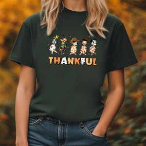 May include: Dark green t-shirt featuring a graphic of cartoon characters in Thanksgiving attire. The word "THANKFUL" is printed in orange and brown. The shirt is made of a soft material.
