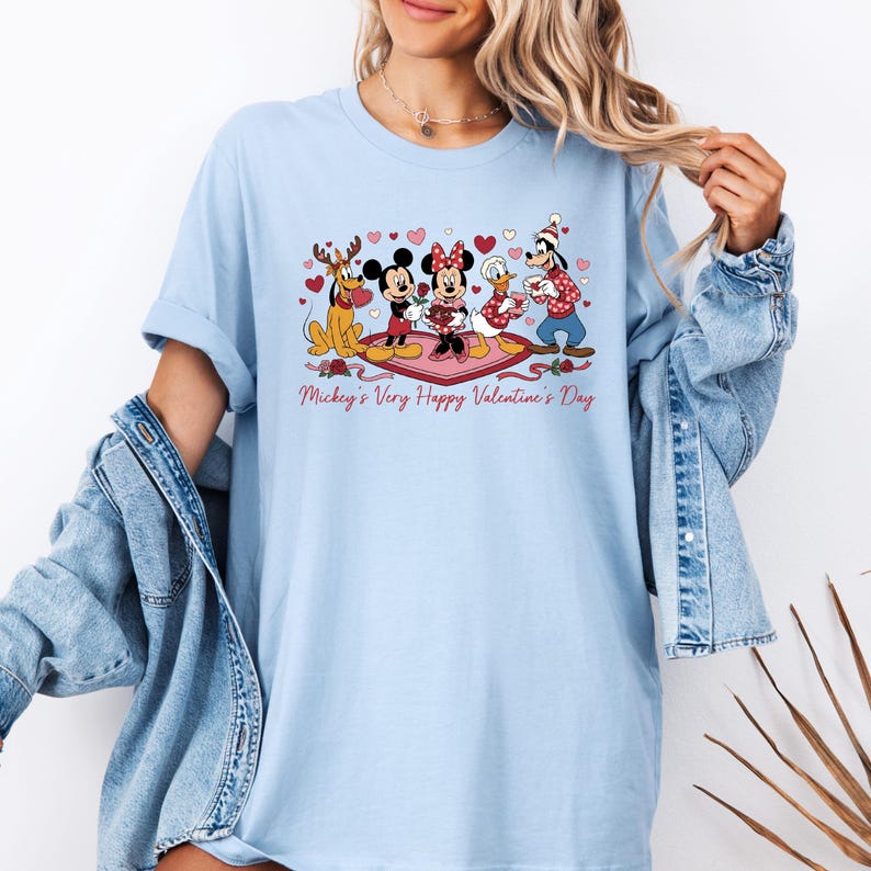 May include: Light blue t-shirt featuring a Valentine's Day design with Mickey Mouse, Minnie Mouse, Donald Duck, Goofy, and hearts. The text "Mickey's Very Happy Valentine's Day" is printed below the characters. The shirt has short sleeves.