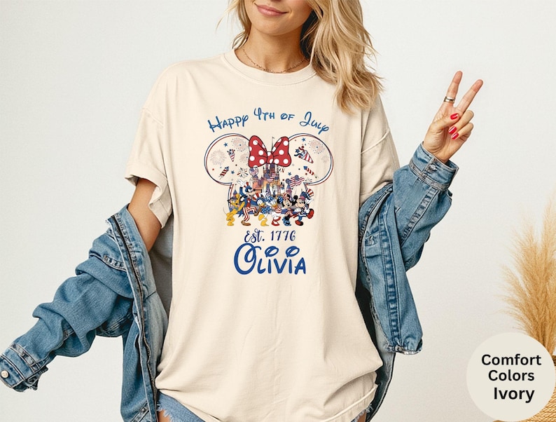 Pode incluir: Camiseta cor marfim com um design patri&oacute;tico com orelhas do Mickey Mouse, um la&ccedil;o vermelho e o texto "Happy 4th of July". O design inclui personagens de desenhos animados e o nome "Olivia" em azul.
