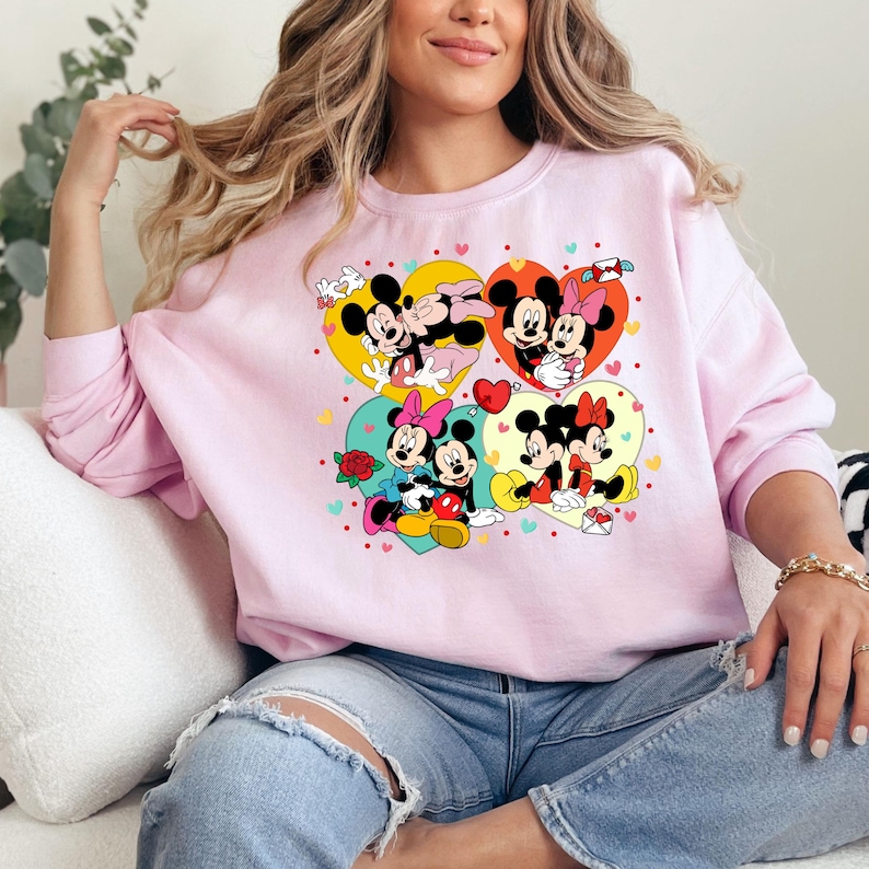 May include: Pink sweatshirt with a graphic print of Mickey Mouse and Minnie Mouse in various poses. The graphic features hearts, flowers, and other romantic details.