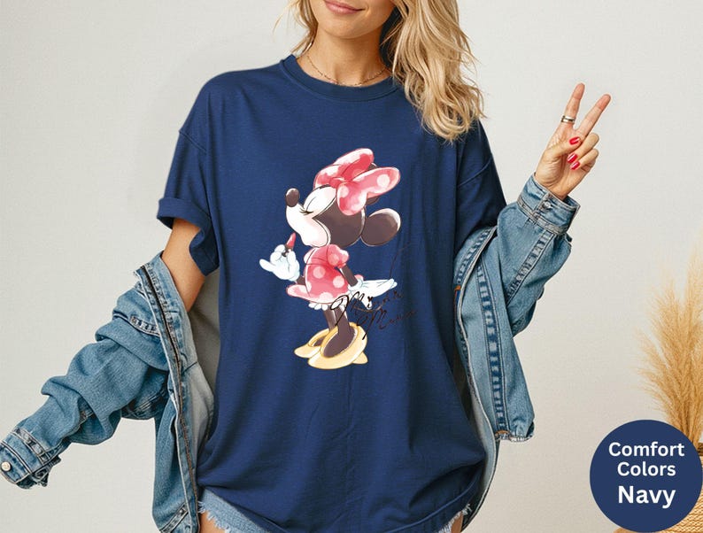 K&ouml;nnte beinhalten: Marineblaues T-Shirt mit Minnie Mouse-Grafik. Die Zeichentrickfigur tr&auml;gt eine rote Schleife und ein gepunktetes Kleid. Das Shirt ist Teil der Comfort Colors-Kollektion.