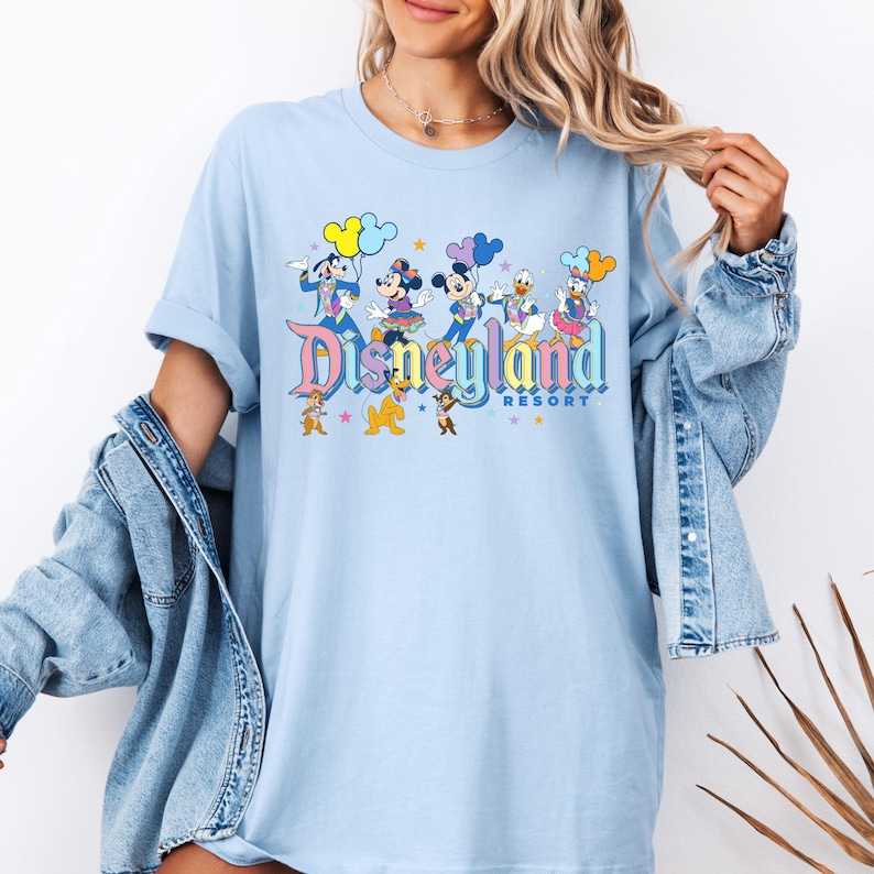 May include: Light blue t-shirt featuring a graphic print of Mickey Mouse, Minnie Mouse, Donald Duck, and Goofy. The graphic includes the text "Disneyland Resort" in a rainbow font.