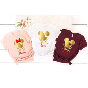 May include: Three white t-shirts with a gold Mickey Mouse head and crossed swords design. The shirts are personalized with the names "Mommy", "Ethan", and "Disney".