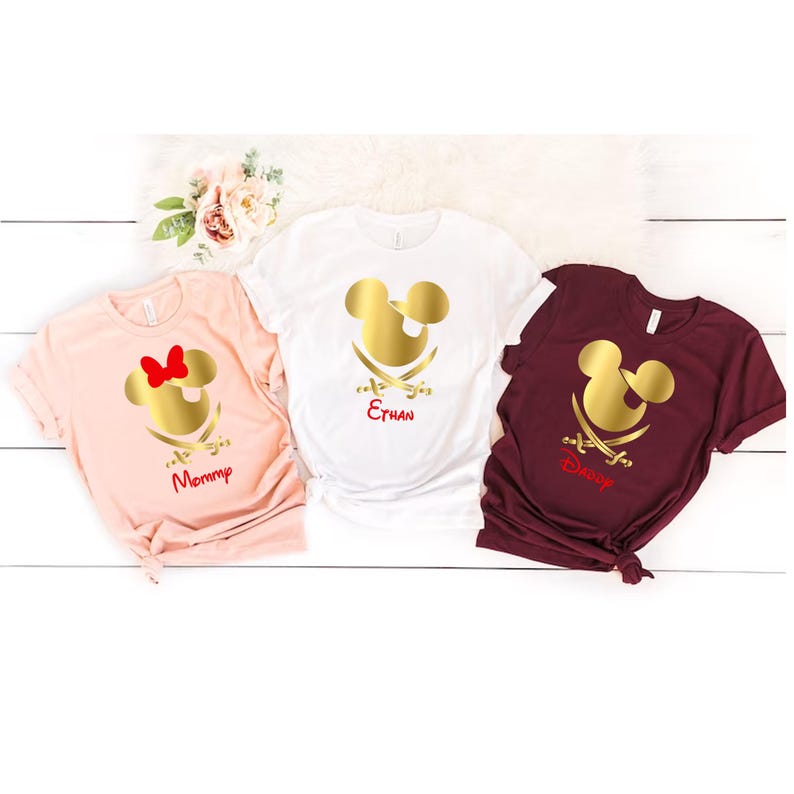 May include: Three white t-shirts with a gold Mickey Mouse head and crossed swords design. The shirts are personalized with the names "Mommy", "Ethan", and "Disney".
