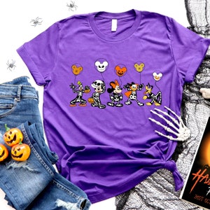 Op de afbeelding: Paars T-shirt met een afbeelding van Mickey Mouse, Minnie Mouse, Donald Duck, Goofy en Pluto verkleed als skeletten die Halloween-ballonnen vasthouden.