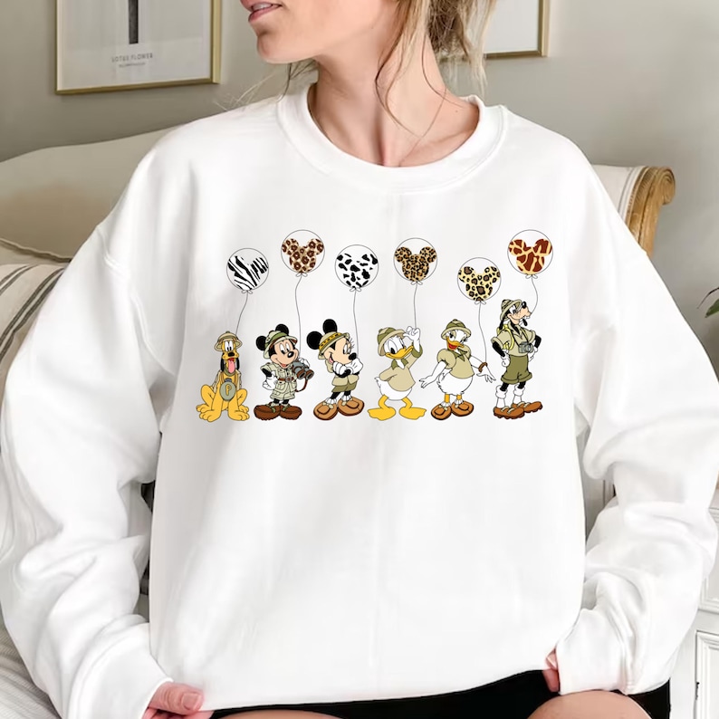 Pode incluir: Camisola branca com um gr&aacute;fico de Mickey Mouse, Donald Duck, Pateta e Pluto segurando bal&otilde;es de estampa animal. Os personagens est&atilde;o usando roupas de saf&aacute;ri.