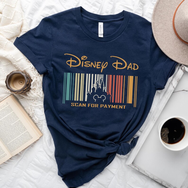 May include: A navy blue t-shirt with a vintage-style graphic that says "Disney Dad" with a barcode underneath that features a silhouette of Cinderella's castle and a Mickey Mouse head. The text below the barcode says "Scan for Payment".