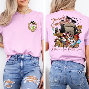 May include: Pink t-shirt with a cartoon pirate character on the front and a graphic of Mickey Mouse and friends on a pirate ship on the back. The back graphic includes the text "Pirates of the Caribbean" and "A Pirate's Life for Me Lyrics".