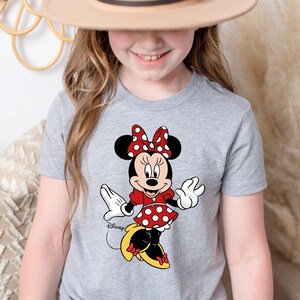K&ouml;nnte beinhalten: Ein graues T-Shirt mit einem roten und wei&szlig;en Polka-Dot-Minnie-Maus-Motiv. Das Motiv zeigt Minnie Maus in einem roten Kleid mit wei&szlig;en Punkten, gelben Schuhen und einer roten Schleife mit wei&szlig;en Punkten. Der Text "Disney" ist in Schwarz unter dem Motiv gedruckt.