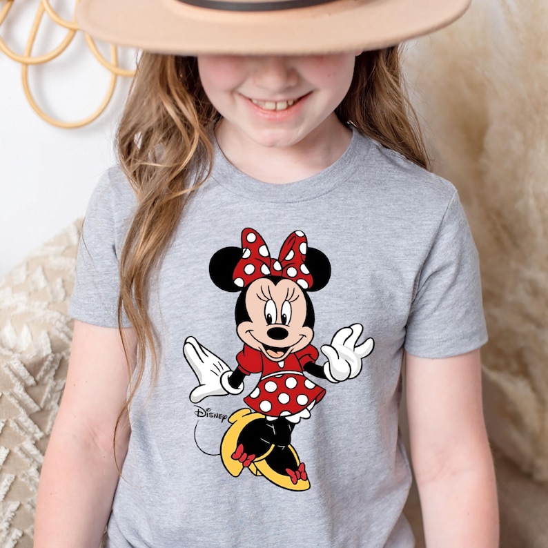 K&ouml;nnte beinhalten: Ein graues T-Shirt mit einem roten und wei&szlig;en Polka-Dot-Minnie-Maus-Motiv. Das Motiv zeigt Minnie Maus in einem roten Kleid mit wei&szlig;en Punkten, gelben Schuhen und einer roten Schleife mit wei&szlig;en Punkten. Der Text "Disney" ist in Schwarz unter dem Motiv gedruckt.
