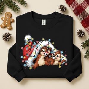 May include: Black sweatshirt featuring Chip and Dale characters with a Santa hat and Christmas lights. The design includes a gingerbread man, pine cones, and evergreen branches, creating a festive holiday theme.