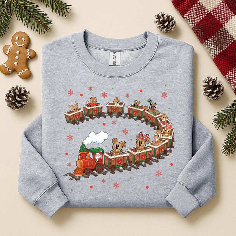 May include: A light gray sweatshirt featuring a festive train design. The train is composed of gingerbread-style cars, each carrying a different character. The train engine is red and green, with a puff of white smoke. Red and green snowflakes are scattered around the train.
