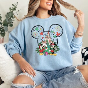 Puede incluir: Sudadera azul claro con un dise&ntilde;o de Mickey Mouse con tem&aacute;tica navide&ntilde;a. Mickey y Minnie est&aacute;n decorando un &aacute;rbol de Navidad frente al Castillo de Cenicienta. El dise&ntilde;o incluye fuegos artificiales y corazones.