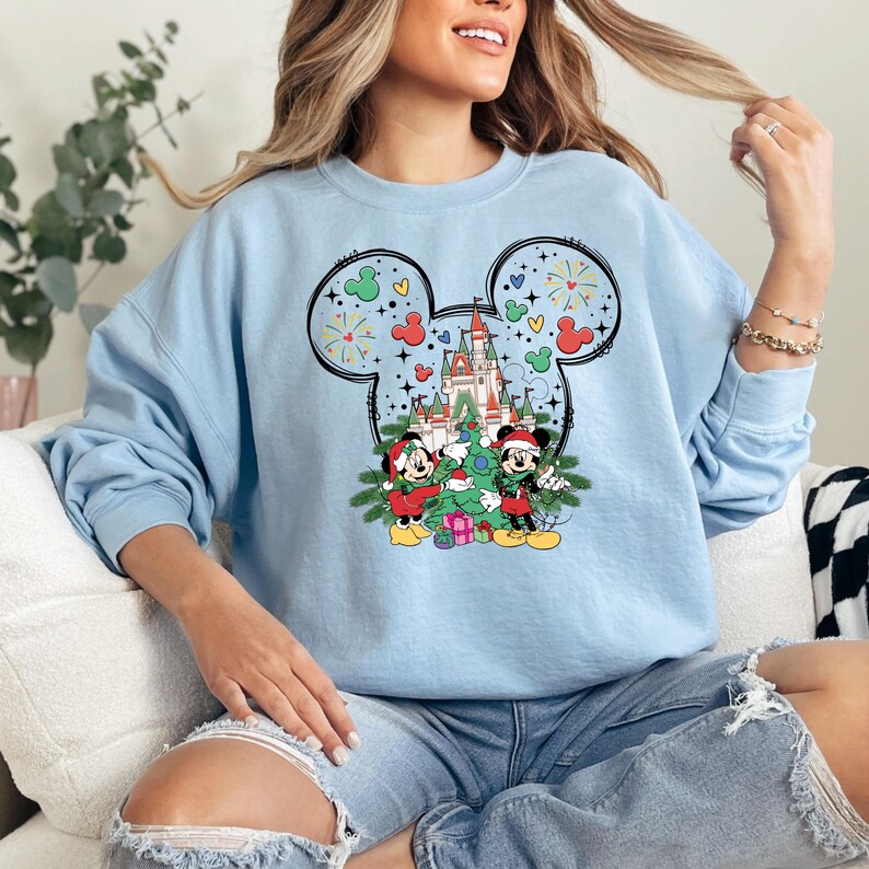 Puede incluir: Sudadera azul claro con un dise&ntilde;o de Mickey Mouse con tem&aacute;tica navide&ntilde;a. Mickey y Minnie est&aacute;n decorando un &aacute;rbol de Navidad frente al Castillo de Cenicienta. El dise&ntilde;o incluye fuegos artificiales y corazones.