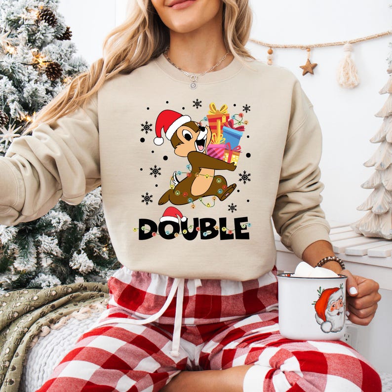 Puede incluir: Una sudadera de Navidad beige con una imagen de dibujos animados de Chipmunk llevando regalos y la palabra "DOUBLE" impresa en la parte delantera. La sudadera la lleva puesta una persona que lleva pantalones de pijama a cuadros rojos y blancos.