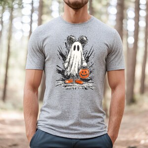 Peut inclure: T-shirt gris avec un motif d'Halloween. Le motif repr&eacute;sente un fant&ocirc;me avec des oreilles de Mickey Mouse, tenant un seau de citrouille et portant des chaussures orange. Le motif est plac&eacute; sur un fond d'&eacute;claboussures de peinture noire.