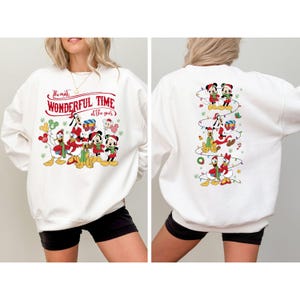 May include: White sweatshirt featuring a festive design with Disney characters. The front displays Mickey, Minnie, Donald, and Goofy with the text "The Most Wonderful Time of the Year." The back shows the characters in a vertical Christmas lights design.