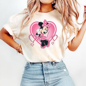 May include: A white t-shirt with a pink heart design featuring Minnie Mouse in a pink dress with white polka dots. Minnie Mouse is holding a pink bow and has a pink bow in her hair.
