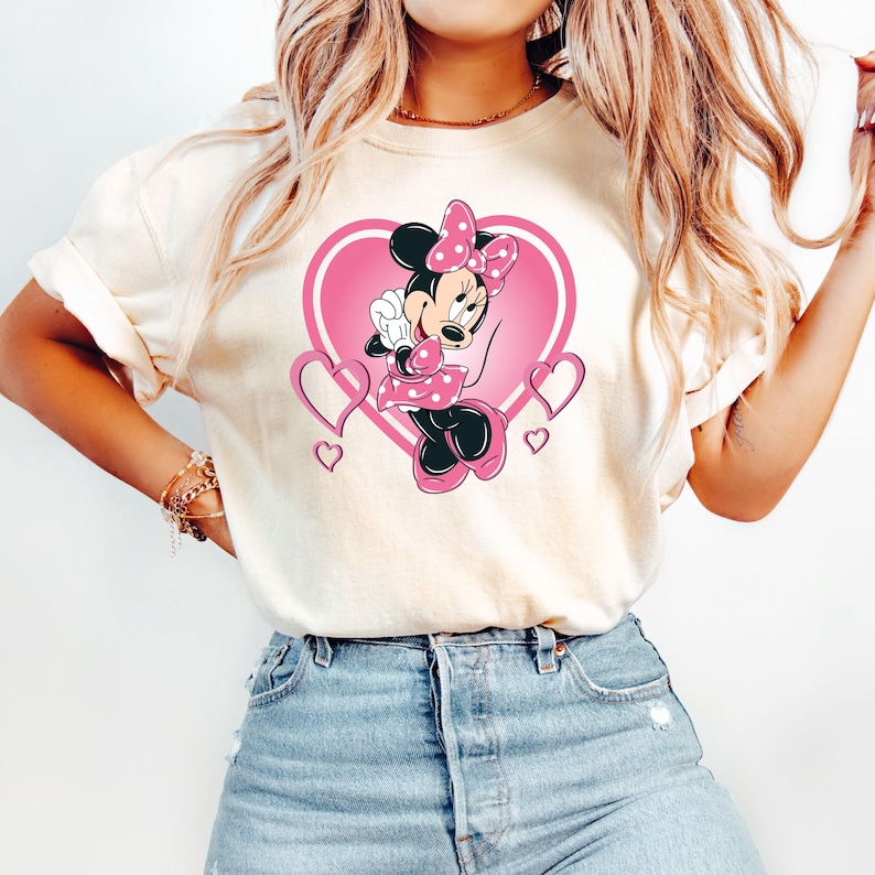 May include: A white t-shirt with a pink heart design featuring Minnie Mouse in a pink dress with white polka dots. Minnie Mouse is holding a pink bow and has a pink bow in her hair.