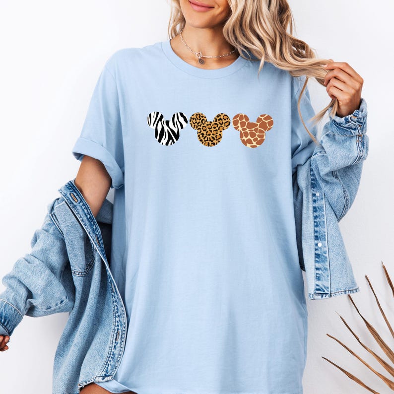 May include: Light blue t-shirt featuring three Mickey Mouse head silhouettes with animal print patterns: zebra, leopard, and giraffe. The shirt has a relaxed fit and short sleeves. The model is wearing a denim jacket.