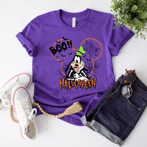 Puede incluir: Camiseta morada con una imagen de dibujos animados de Goofy vestido de esqueleto con el texto "BOO!" y "HALLOWEEN" en ella.