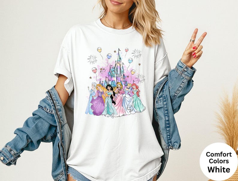 K&ouml;nnte beinhalten: Wei&szlig;es T-Shirt mit einer farbenfrohen Illustration von Prinzessinnen vor einem Schloss. Das Design umfasst rosa, blaue und lila Akzente mit Feuerwerk und Luftballons. Das Shirt ist mit "Comfort Colors White" gekennzeichnet.