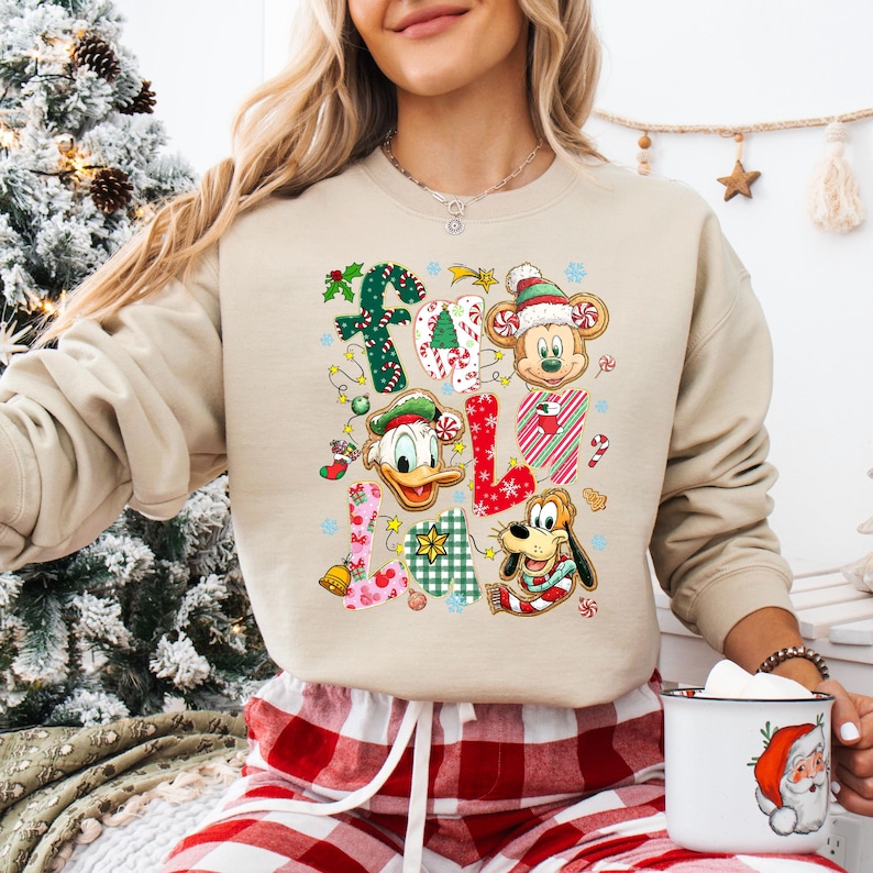 K&ouml;nnte beinhalten: Beiges Sweatshirt mit einem farbenfrohen Weihnachtsdesign mit Micky Maus, Donald Duck und Pluto. Der Text "FALALA" ist mit festlichen Mustern und Weihnachtselementen in das Design integriert. Ein gem&uuml;tliches und festliches Kleidungsst&uuml;ck.